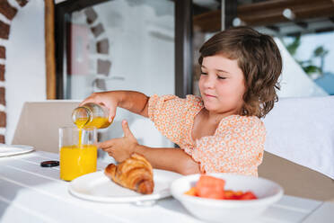 Braunhaariges Kind sitzt am Tisch und gießt Orangensaft in ein Glas beim Frühstück mit Croissant und Obstsalat im Café - ADSF41132