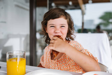 Cheerful preschool girl eating croissant and looking at camera while sitting at table with glass of orange juice in cafe - ADSF41133