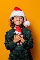 Cute girl in green dress and Santa hat standing looking at camera with crochet toy during Christmas celebration against yellow background - ADSF41136