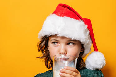 Niedliches Mädchen mit Weihnachtsmannmütze nippt an Milch aus einem Glas und schaut weg während der Weihnachtsfeier vor gelbem Hintergrund - ADSF41139