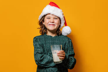 Nettes fröhliches Mädchen mit Weihnachtsmannmütze nippt Milch aus einem Glas und schaut in die Kamera während der Weihnachtsfeier vor gelbem Hintergrund - ADSF41140