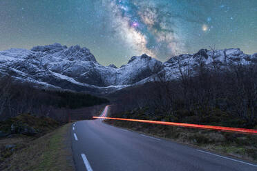 Breathtaking scenery of rocky mountain range covered with snow and empty asphalt road under night sky glowing stars of Milky Way in Norway - ADSF41168