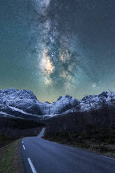Breathtaking scenery of rocky mountain range covered with snow and empty asphalt road under glowing stars of Milky Way in night sky in Norway - ADSF41169