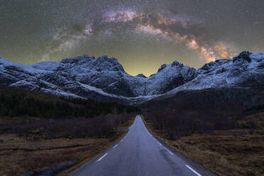 Breathtaking scenery of rocky mountain range covered with snow and empty asphalt road under glowing stars of Milky Way in night sky in Norway - ADSF41170