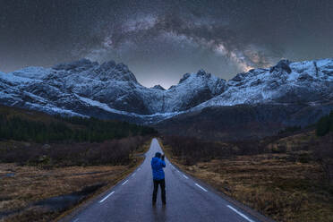 Fernansicht eines Forschers, der auf einer asphaltierten Straße in den verschneiten Bergen unter dem nächtlichen Sternenhimmel der Milchstraße in Norwegen steht - ADSF41174