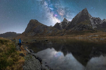 Remote view of unrecognizable traveling photographer taking picture of night sky glowing stars of Milky Way in Lofoten Islands, Norway - ADSF41175