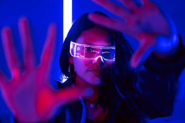 Ethnic female touching air while experiencing virtual reality in modern transparent VR glasses in dark studio with bright neon illumination looking at camera - ADSF41231