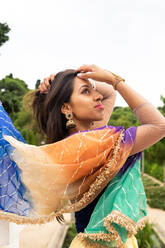 Seitenansicht einer jungen ethnischen indischen Frau in einem stilvollen traditionellen Sari und Schmuck, die in einem grünen Park steht und wegschaut - ADSF41233