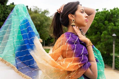 Side view of arrogant young ethnic woman in traditional sari standing in park among green trees and holding hair while showing big earring - ADSF41234