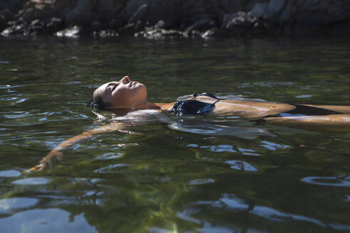 Ruhige Frau im schwarzen Bikini schwimmt im klaren Wasser des Bergflusses und entspannt sich mit geschlossenen Augen - ADSF41239