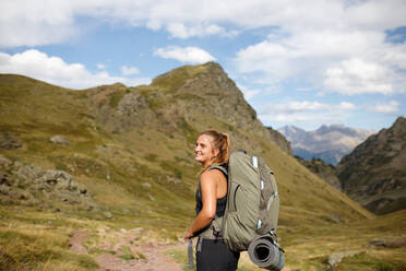 Side view of happy young woman with backpack and mat smiling and looking away against green mountains and cloudy blue sky - ADSF41242