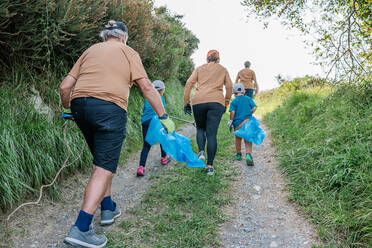 Back view of unrecognizable people with kids with plastic bags walking on grassy path during environmental campaign in countryside - ADSF41247