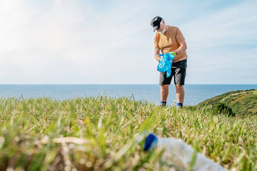 Empty used plastic bottle on grass with senior volunteer collecting garbage in background during environmental campaign in countryside - ADSF41252