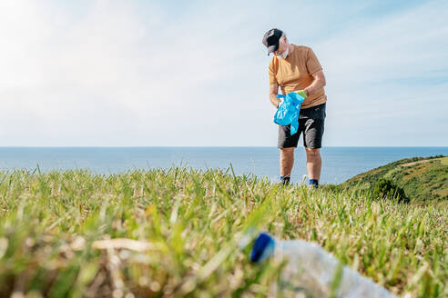 Leere gebrauchte Plastikflasche auf Gras mit einem älteren Freiwilligen, der im Hintergrund während einer Umweltkampagne auf dem Lande Müll sammelt - ADSF41252