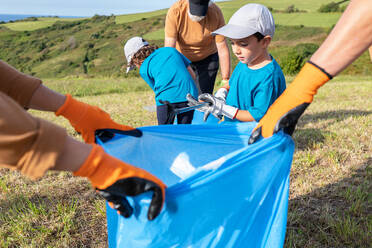 Eco friendly family in caps picking garbage via thrash tongs into blue bin bag for recycling on grassy hill near sea - ADSF41258