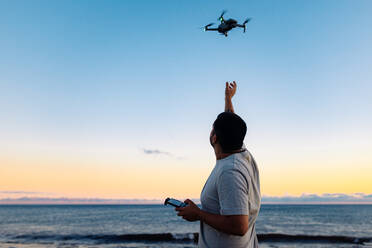 Unrecognizable black male in casual clothes launching modern drone against sunset sky while spending time on beach near waving sea - ADSF41267
