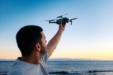 Unrecognizable black male in casual clothes launching modern drone against sunset sky while spending time on beach near waving sea - ADSF41269
