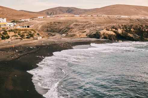 Von oben malerische Szenerie des mächtigen Ozeans mit schäumenden Wellen am Strand mit vulkanischem schwarzem Sand, umgeben von Hügeln bei Sonnenuntergang auf Fuerteventura - ADSF41273