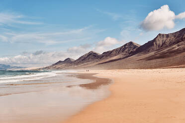 In der Ferne unerkennbare Menschen, die vor der atemberaubenden Kulisse der schäumenden Wellen des Ozeans spazieren gehen, die an der breiten Sandküste in der Nähe von felsigen Bergen mit scharfen Gipfeln gegen den strahlend blauen Himmel auf Fuerteventura rollen - ADSF41276