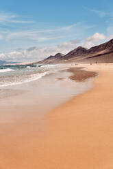 In distance unrecognizable people walking on breathtaking scenery of foamy waves of ocean rolling on wide sandy coast near rocky mountains with sharp peaks against bright blue sky in Fuerteventura - ADSF41277