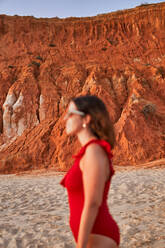 Seitenansicht einer Frau in roter Unterwäsche und Sonnenbrille am Sandstrand von Falesia in der Nähe von Sandsteinklippen bei Sonnenuntergang in der Algarve, Portugal - ADSF41278