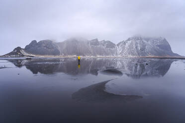 Distant view of unrecognizable traveler standing on wet black sand beach near waving sea against mountain ridge and cloudy sky in Iceland - ADSF41294