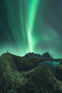 Unbekannter Fotograf mit Fotokamera, der auf einer Bergklippe steht und den Blick auf das grüne Polarlicht einfängt, das den dunklen Nachthimmel auf den Lofoten beleuchtet - ADSF41301
