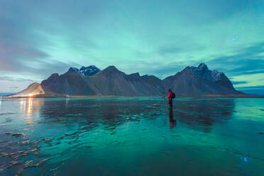Unrecognizable traveler photographer with photo camera standing on frozen lake in rocky snowy mountains and admiring view of green aurora borealis illuminating dark night sky in Lofoten Islands - ADSF41307