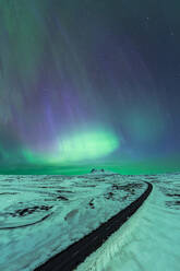 Breathtaking scenery of empty curvy asphalt road going amidst field covered with snow under night sky with green aurora borealis in Iceland - ADSF41320