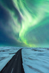 Breathtaking scenery of empty curvy asphalt road going amidst field covered with snow under night sky with green aurora borealis in Iceland - ADSF41321