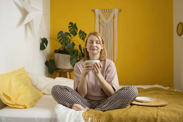 Full Body of lächelnd Ingwer barfuß Dame in gestreiften Hosen sitzen auf bequemen Bett und genießen Morgenkaffee gegen orange Wand mit Makramee Dekoration und grüne Monstera, während wegschauen - ADSF41344