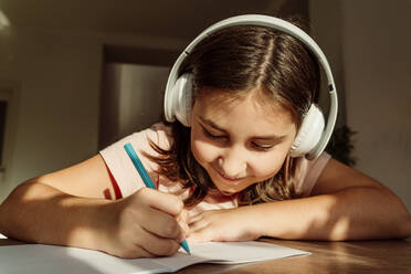 Smiling girl wearing wireless headphones writing in book at desk - OSF01150