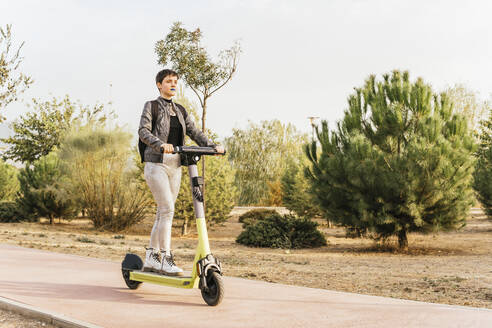 Woman riding electric push scooter on footpath - MGRF00838