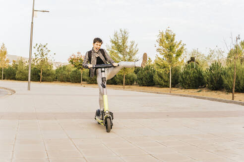 Playful woman riding electric push scooter with leg up - MGRF00841