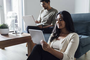 Junge Frau lernt mit Tablet-PC und Mann im Hintergrund zu Hause - MEUF08609