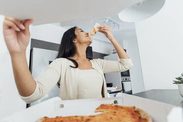 Glückliche junge Frau isst Pizza zu Hause - MEUF08624