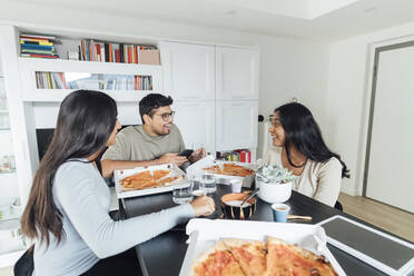 Glückliche Frauen und Männer essen Pizza am Esstisch im Wohnzimmer - MEUF08625