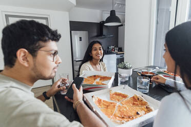 Mann mit Handy in der Hand sitzt mit Frauen zusammen und isst Pizza zu Hause - MEUF08626