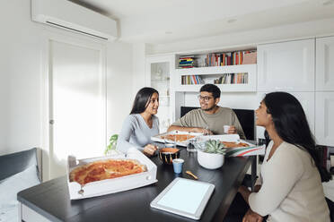 Glückliche Frauen und Männer diskutieren und essen Pizza am Esstisch im Wohnzimmer - MEUF08627