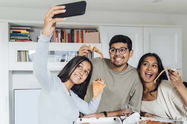 Lächelnde Frau nimmt Selfie durch Smartphone mit Frau und Mann zeigt Pizzastück zu Hause - MEUF08631