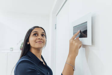 Smiling woman using smart home device on wall - MEUF08641