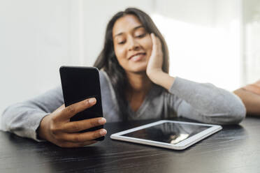 Lächelnde Frau, die ein Smartphone benutzt und mit einem Tablet-PC auf einem Tisch zu Hause sitzt - MEUF08652