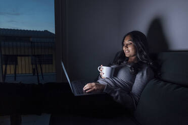 Smiling young woman holding coffee cup using laptop at home - MEUF08697