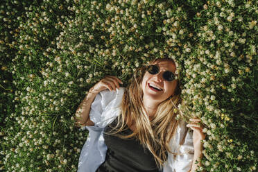 Happy woman wearing sunglasses lying on meadow of clover flowers - MDOF00296