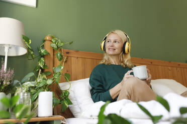 Nachdenkliche Frau beim Musikhören mit einer Tasse Tee auf dem Bett - TYF00451