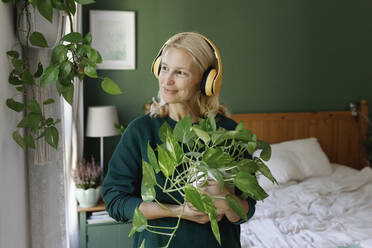 Woman with headphones listening to music carrying green plant in bedroom - TYF00460