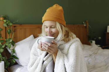 Lächelnde Frau mit Strickmütze, die im Schlafzimmer an Tee riecht - TYF00461