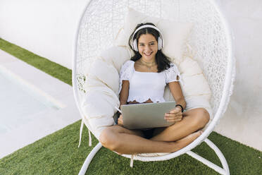 Happy girl wearing headphones sitting with laptop on hanging chair - EGHF00657
