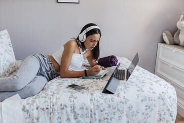 Girl wearing headphones writing notes lying on bed with gadgets at home - EGHF00664