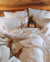 Draufsicht auf einen nicht erkennbaren Mann im weißen Bademantel, der auf einem weichen Bett steht und eine positive junge Freundin fotografiert, die unter einer Decke liegt und zwei Finger zeigt - ADSF41348